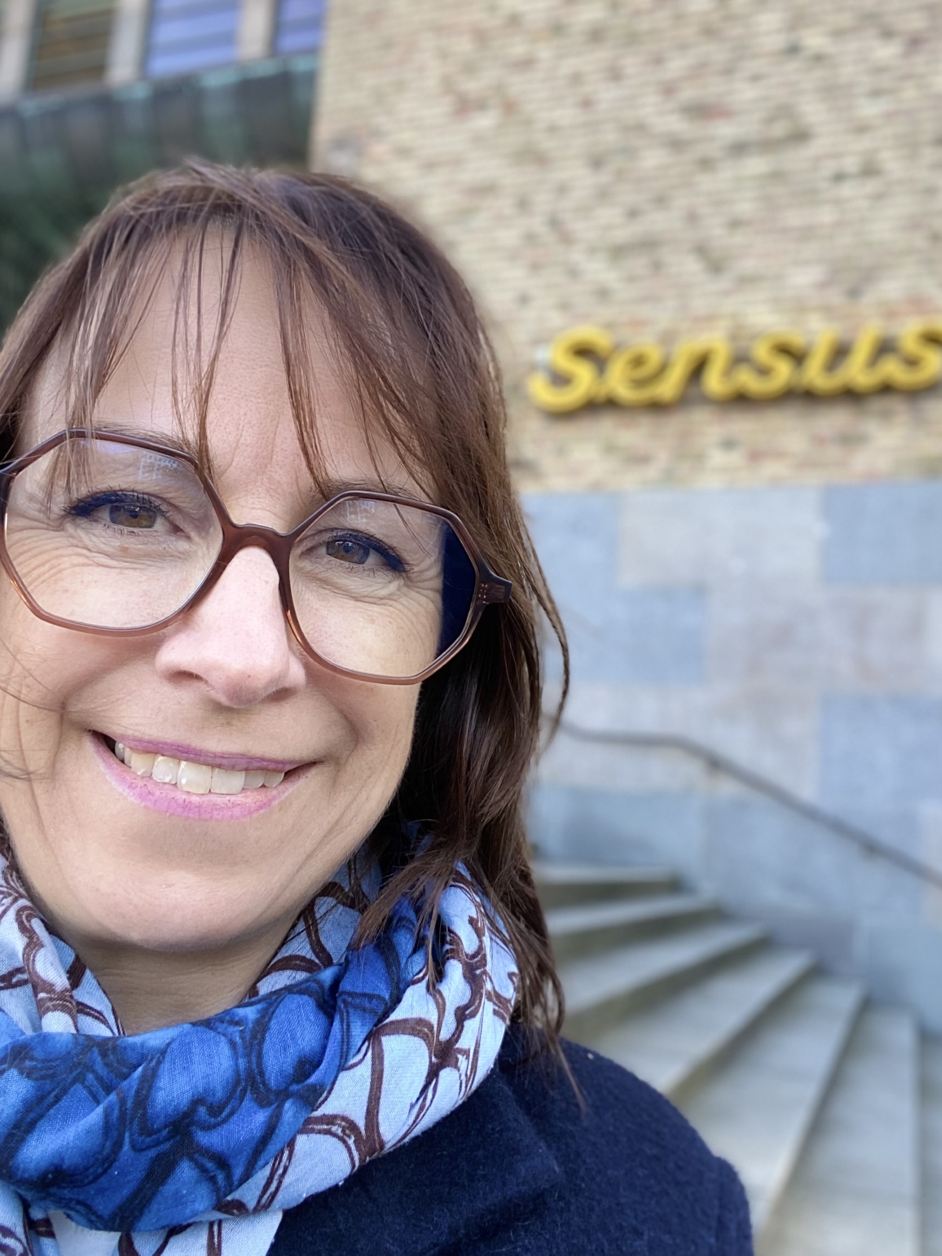 Person står framför en byggnad med en skylt som säger "Sensus". Det är en solig dag.