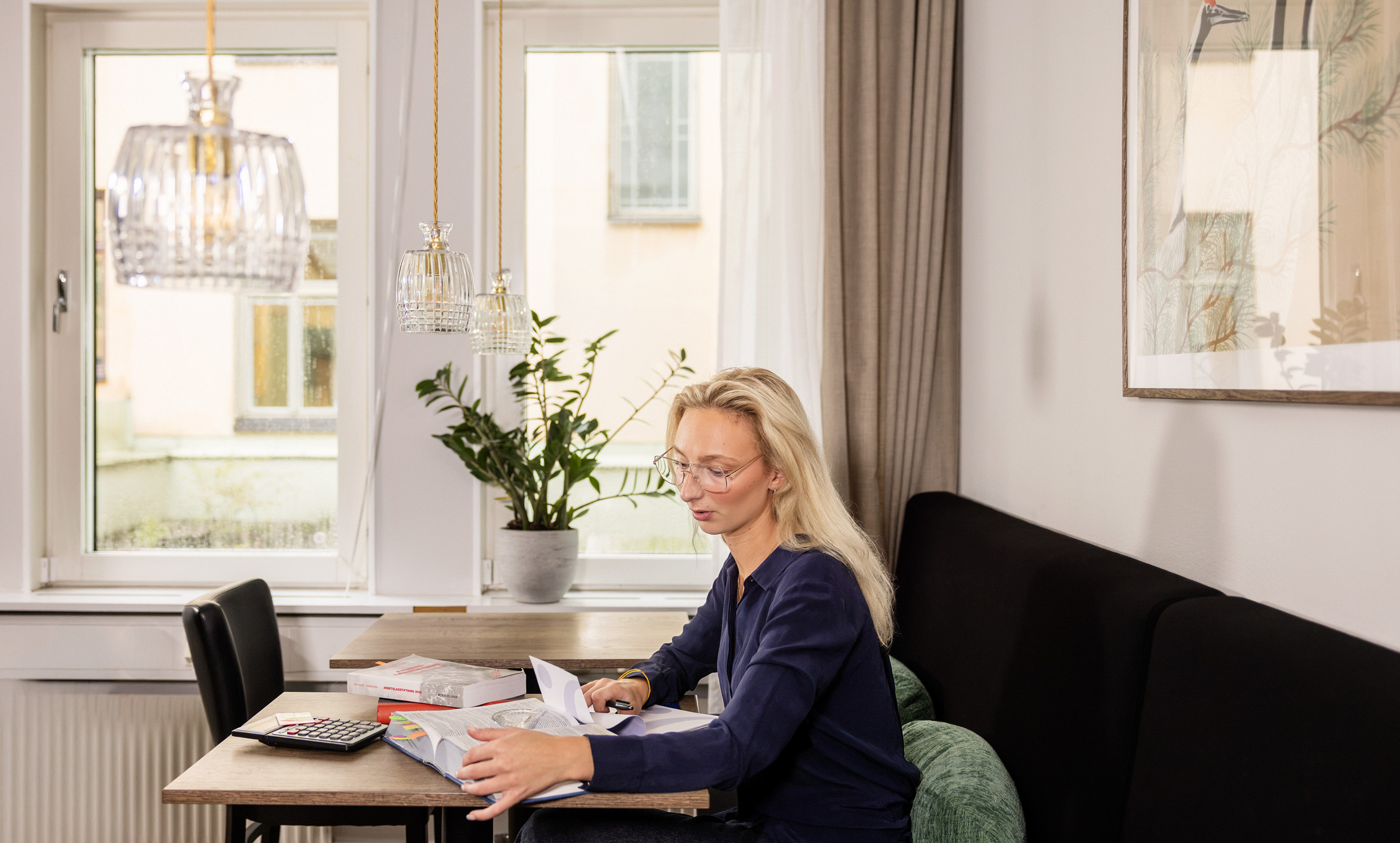 En kvinna sitter vid ett bord och studerar med kalkylator, böcker och papper i ett ljust rum.